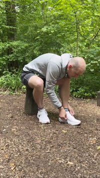 Des baskets de sport blanches avec des accents gris, en matériau synthétique et avec des semelles texturées. Une personne est assise sur une souche en bois en train de lacer ses chaussures.