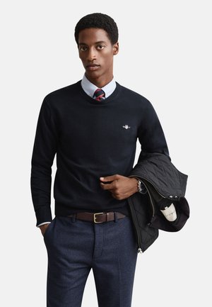 Pull bleu marine à col rond, orné d'un petit logo. Associé à une chemise bleu clair, une cravate à motifs et un pantalon bleu foncé. Tenant une veste noire.