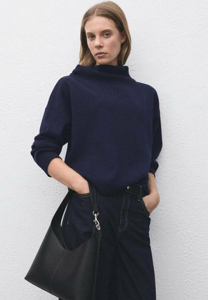 Vrouw in een marineblauwe trui en broek, met een zwarte leren handtas, staande tegen een witte textuurwand.