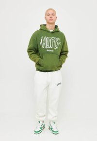 Giovane uomo con i capelli biondi corti che indossa un felpa verde con scritte bianche, pantaloni della tuta bianchi e sneakers bianche con accenti verdi.