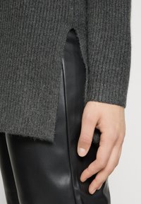 Grauer gerippter Strickpullover mit seitlichem Schlitz, kombiniert mit enganliegenden schwarzen Lederhosen. Hand auf der Hose ruhend, zeigt Textur und Lagen.