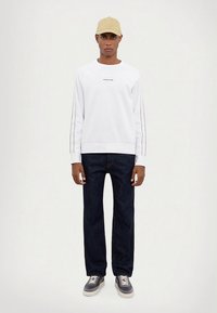 Fehér, fekete oldalsávos környakú pulóver, sötétkék farmerrel és szürke sportcipővel viselve. Egy bézs sapka teszi teljessé a szettet.