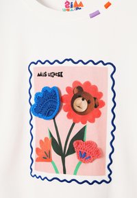 Biała koszulka z kolorową grafiką przedstawiającą niedźwiedzia w kompozycji kwiatowej, obramowana niebieską taśmą falistą i ozdobiona fakturowanymi elementami szydełkowymi.