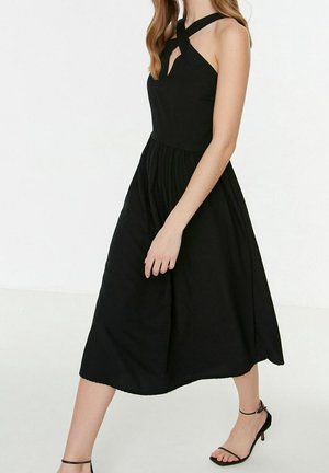 Robe noire sans manches avec une ouverture en forme de goutte et des bretelles entrecroisées, jupe fluide et tissu doux, longueur au-dessus de la cheville portée avec des sandales plates.