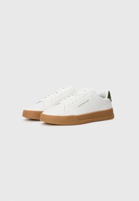 Para białych niskich sneakersów z brązowymi podeszwami, białymi sznurówkami, zielonym paskiem na pięcie oraz małym logo Tommy Hilfiger z boku.