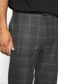 Grå rutig byxor med flat front, med blå och bruna linjer, tillverkade av en texturerad tyg. Sidofickor synliga.