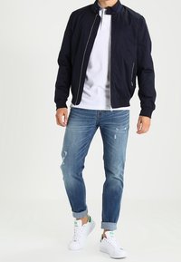 Marinblå bomberjacka, vit polo, slitna blå jeans och vita sneakers med gröna detaljer. Jackan har en dragkedja och sidofickor.