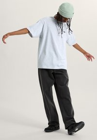 Šedé oversized tričko, černé volné kalhoty a černé sneakers. Model nosí světle zelenou pletenou čepici s texturovaným vzorem.