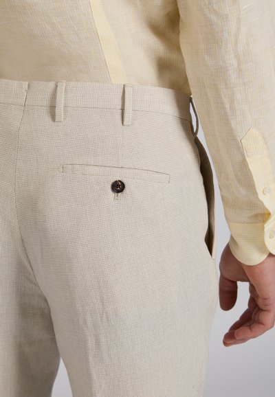 Pantalon beige texturé avec une poche arrière boutonnée, porté avec une chemise à manches longues jaune clair, vue du côté droit avec la main visible.