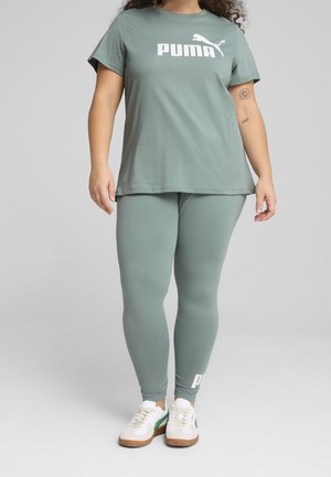 Vrouw draagt een lichtgroen Puma T-shirt en legging met witte sneakers, staand tegen een eenvoudige grijze achtergrond.