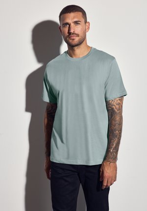Lys teal kortærmet t-shirt lavet af glat stof, med en crew neck, iført af en person med tatoverede arme mod en ensfarvet baggrund.