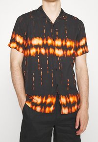 Korte mouw blouse van zwarte stof met een levendig oranje en geel tie-dye patroon, voorzien van een knoopsluiting aan de voorkant en een camp-kraag ontwerp.