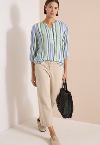 Femme portant un chemisier rayé bleu, vert et blanc avec un pantalon large beige, tenant un sac à main tissé noir, marchant à l'intérieur.