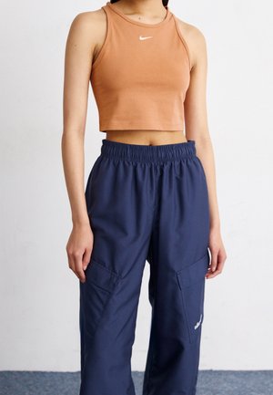 Kobieta w bezrękawnym, krótkim brązowym topie Nike i luźnych granatowych spodniach typu cargo stojąca na tle białego tła.