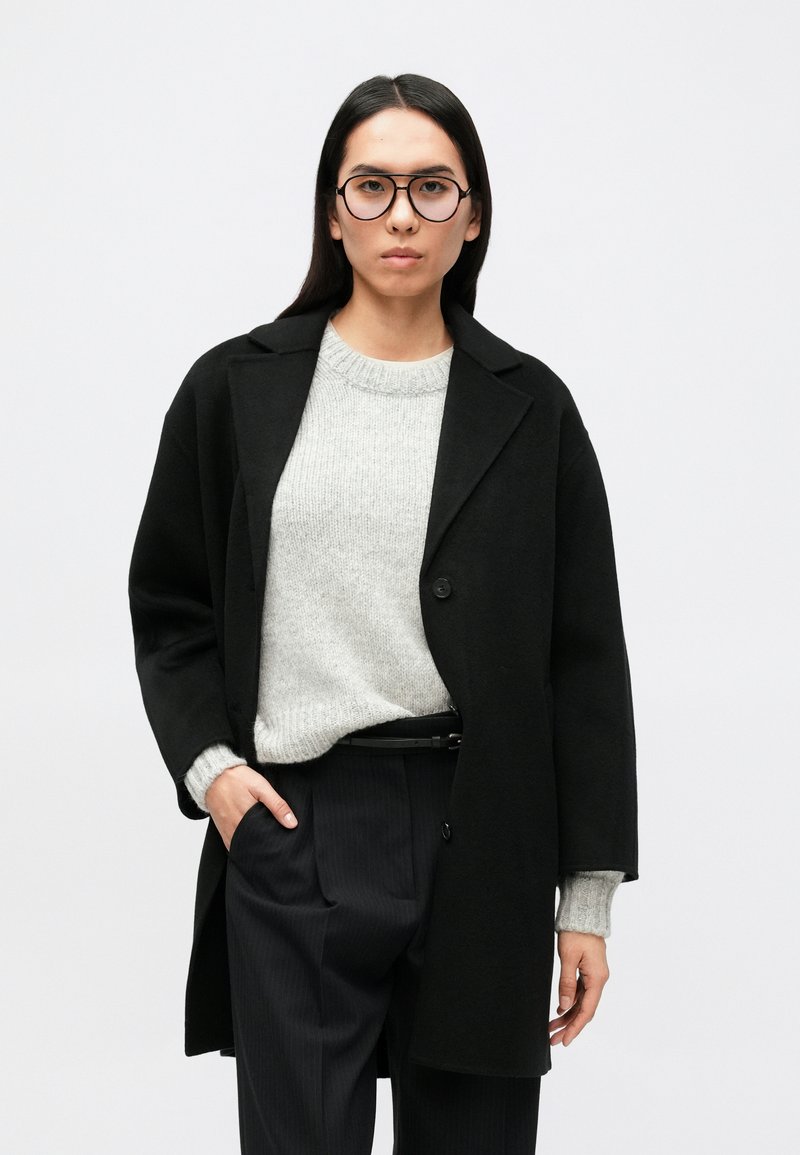 Čierny oversize kabát s vypálenými klopami, nosený cez svetlosivý pletený sveter, doplnený tmavými pruhovanými nohavicami. Jednoduché zapínanie na gombíky.