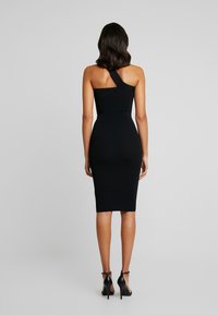 Robe noire moulante à une épaule, en tissu lisse, arrivant au milieu du mollet. Portée avec des talons noirs à brides.