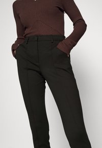 Personne portant un pantalon noir sur mesure avec poches latérales et un haut marron côtelé à manches longues, mains partiellement dans les poches, sur fond uni.