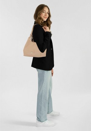 Borsa a mano beige chiaro con una texture liscia, caratterizzata da un logo argentato e una tracolla regolabile, tenuta da una persona con una giacca nera e jeans chiari.