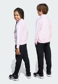 Svijetlopinkasta jakna sa zipom i bijelim prugama, uparen s crnim trenirkama i crnim tenisicama s bijelim detaljima.