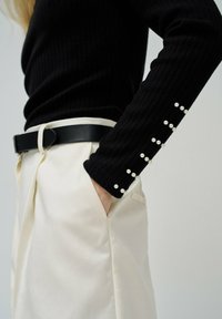 Zwarte geribde longsleeve top met parel detail op de manchetten, gecombineerd met crèmekleurige broek en een zwarte riem. De stof lijkt zacht en textuur te hebben.