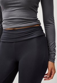 Langarm, aansluitend grijs topje met geplooide textuur bij de taille, gecombineerd met hoge zwarte legging, beide gemaakt van rekbare stof.