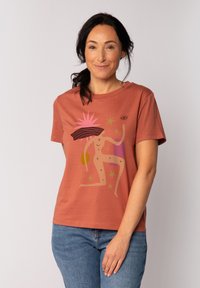 T-shirt en coton couleur rouille avec un design graphique d'une femme entourée d'éléments abstraits en rose, jaune et vert, à manches courtes et à coupe décontractée.