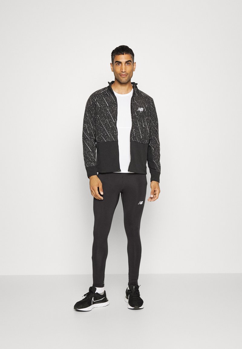 Schwarz-weiß gemusterte Zip-Jacke mit elastischen Bündchen, kombiniert mit schwarzen Leggings und schwarzen Sportschuhen, die ein Logo am Bein aufweisen.
