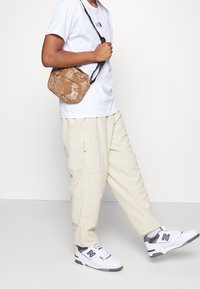 Bolso cruzado de estampado marrón con una correa negra ajustable, llevado con una camisa blanca y pantalones beige. Unos sneakers blancos y grises completan el look.