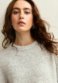 Jongeman met golvend bruin haar die zilveren oorbellen draagt en een zachte grijze gebreide trui, kijkend recht vooruit met een neutrale uitdrukking.