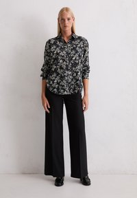 Femme avec des cheveux blonds raides portant une chemise noire à fleurs à boutons, un pantalon large noir et des mocassins noirs, se tenant contre un mur blanc.