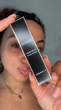 Personne avec un gloss transparent et un bandeau tient une boîte de highlighter MAC Skinfinish Lightstruck près de son visage.