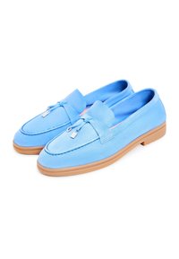 Mocassins en daim bleu clair avec détails à pampilles et semelles en caoutchouc marron clair, présentés sous un angle trois quarts sur fond blanc.