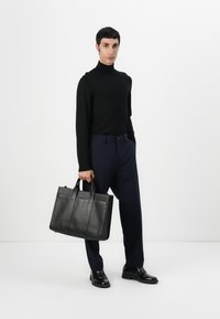 Sort læder tote bag med struktureret form, dobbelt hanke i toppen, tekstureret overflade og lodrette sømme. Model iført en sort turtleneck og marineblå bukser.