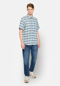 Lyseblå plaid kortærmet skjorte, knap lukket, parret med blå jeans og beige træningssko. Modellen står med hænderne ved siden af.