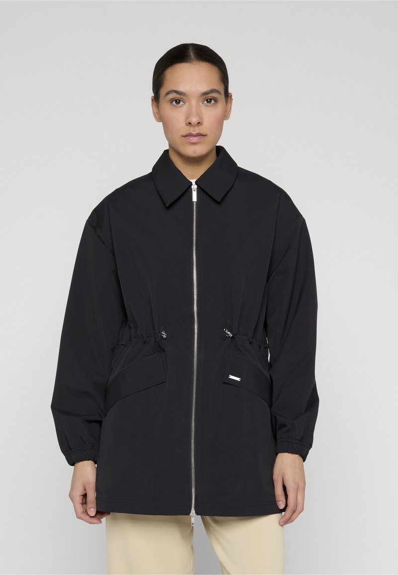 Frau trägt eine schwarze Jacke mit Reißverschluss, Kragen und elastischen Bündchen, kombiniert mit hellbeigen Hosen, und steht vor einem schlichten hellen Hintergrund.