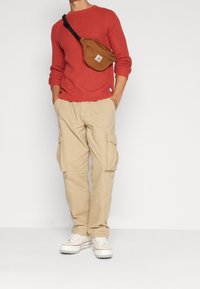 Rød ribstrikket sweater, beige cargobukser med sidelommer og hvide sneakers, accessoriseret med en brun crossbody-taske.