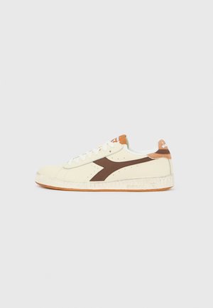 Sneaker crema a basso profilo con logo marrone laterale, lacci bianchi, patch arancione sul tallone e suola traforata su sfondo chiaro semplice.