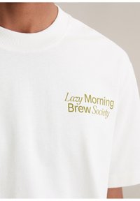 Close-up van een wit T-shirt met "Lazy Morning Brew Society" in gouden letters gedrukt op de linkerkant van de borst.