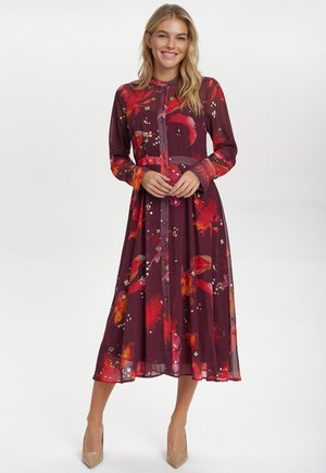 Lungo abito bordeaux con stampa floreale in rosso e giallo, chiusura frontale con bottoni, maniche lunghe e una silhouette fluida. Tessuto morbido e leggero.