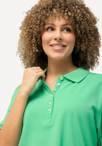 Zelená polo tričko vyrobené z měkké tkaniny s límečkem, vybavené knoflíkovou lištou. Knoflíky mají světlou barvu, která ladí s živou barvou trička.