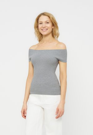 Woman with shoulder-length blonde hair wearing a gray off-shoulder ribbed top and white pants, smiling and standing against a white background.