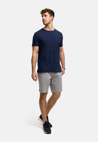 Marineblaues T-Shirt mit kurzen Ärmeln und einer Brusttasche, graue Shorts und marineblaue Sneaker, stehend vor einem einfarbigen Hintergrund.