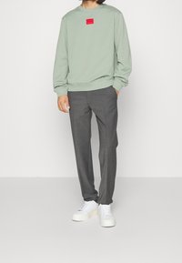 Felpa verde chiaro con patch logo rosso, abbinata a pantaloni grigi e sneakers bianche. Tessuto casual e vestibilità rilassata.