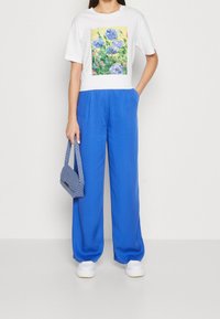 Bílá tričko s květinovým potiskem, modré široké kalhoty a texturovaná modrá kabelka. Bílé tenisky dotvářejí outfit.