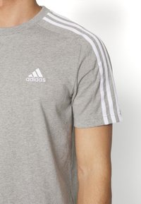 Sivi pamučni majica s okruglim izrezom, koja sadrži tri bijele pruge na svakoj ramenu i izvezeni bijeli Adidas logotip na prsima.