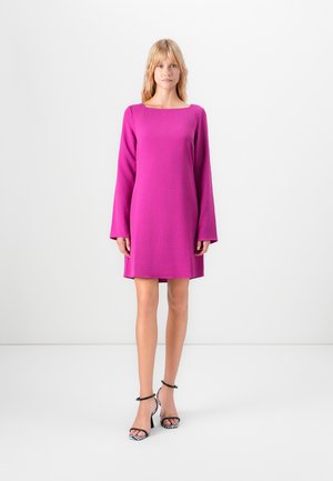 Femme debout dans une robe longue à manches longues rose vif et des sandales à talons hauts rayées noir et blanc devant un fond blanc uni.