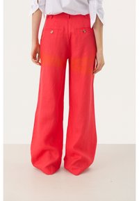 Persoon in hoge taille, wijdvallende rode broek met knoopzakken aan de achterkant en een witte shirt met opgerolde mouwen, staande op een lichte vloer.