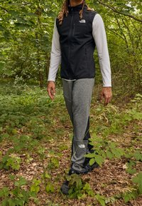 Eine Person geht in einem grünen Wald und trägt schwarze Trailschuhe, graue und schwarze Hose, ein weißes Langarmshirt und eine schwarze ärmellose Weste.