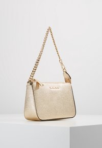 Złota torebka crossbody o teksturze z zamkiem błyskawicznym, łańcuszkowym paskiem oraz kontrastującymi czarnymi wykończeniami. Z przodu znajduje się tłoczone logo.