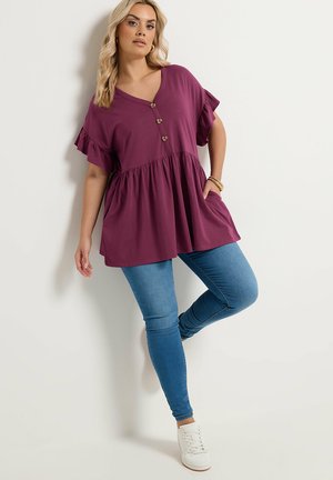 Femme appuyée contre un mur, portant une tunique violette ample avec des boutons, un jean slim bleu, des baskets blanches et des bracelets en or.